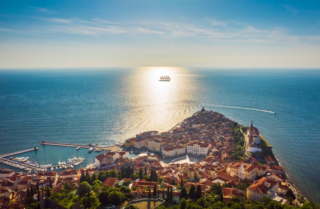 Ein Foto aus der Vogelperspektive von Piran, einer slowenischen Küstenstadt. Im unteren Teil des Bildes ist die Stadt zu sehen und im mittleren/oberen Teil sieht man das Meer mit einem Segelschiff.