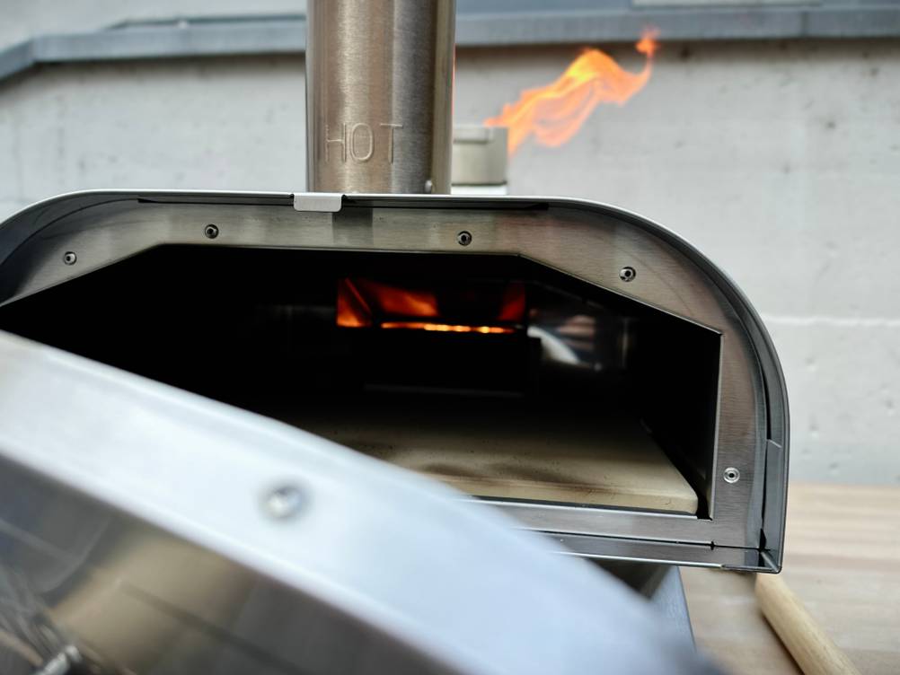 Der Big Horn Outdoors SRPG18003 steht mit aktiver Flamme und heißem Pizzastein auf einem Tisch. Die Flamme ist über dem Kamin sichtbar.