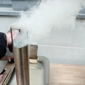 Dichter Rauch steigt aus dem Schornstein des Big-Horn-Outdoors-SRPG18003-Pizzaofens auf. Eine Person beugt sich daneben über den Tisch.