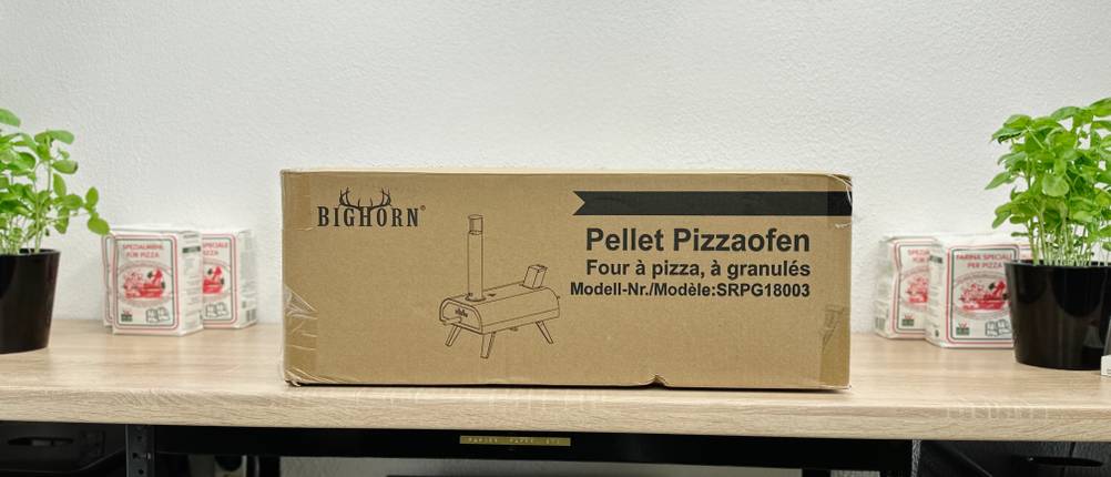 Der Big-Horn-Outdoors-SRPG18003-Pizzaofen in seiner Originalverpackung steht auf einem Tisch zwischen Pizzamehl und Basilikum.