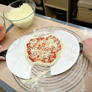 Eine Pizza mit Tomatensauce und Käse wird auf einer Schaufel vorbereitet. Im Hintergrund steht der G3Ferrari G1003210.
