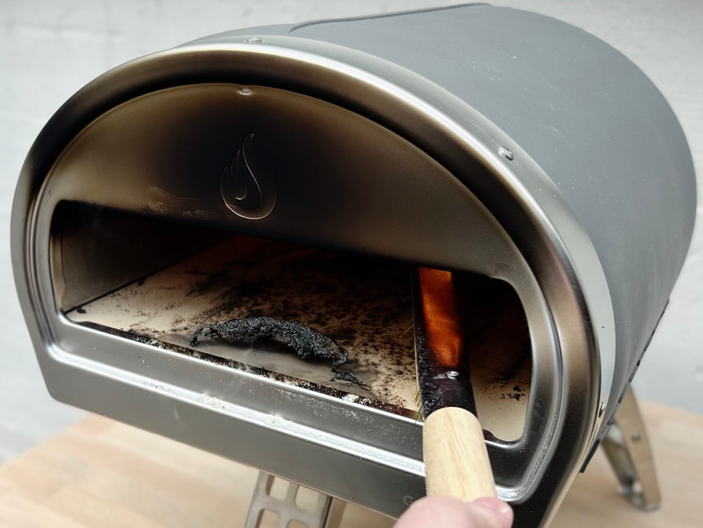 Der Gozney Pizzaofen wird nach dem Backen mit einer Reinigungsbürste gesäubert. Auf dem Stein befinden sich verbrannte Teigreste und Asche.