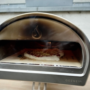 Eine belegte Rohpizza backt im Gozney Pizzaofen auf einem Pizzastein. Rauch steigt leicht aus dem Ofen auf.