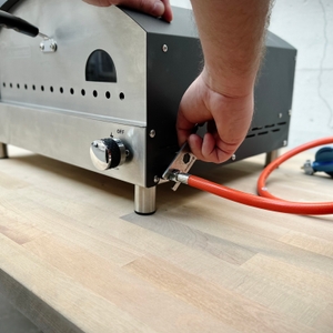 Eine Hand montiert den Gasschlauch am Anschluss des Heidenfeld KY019G Pizzaofens. Der orangefarbene Schlauch ist mit einem Druckminderer verbunden.