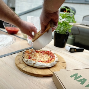 Eine fertige Pizza auf einem Holzbrett wird mit einem Pizzaschneider geteilt. Im Hintergrund stehen Zutaten und Basilikumpflanzen.