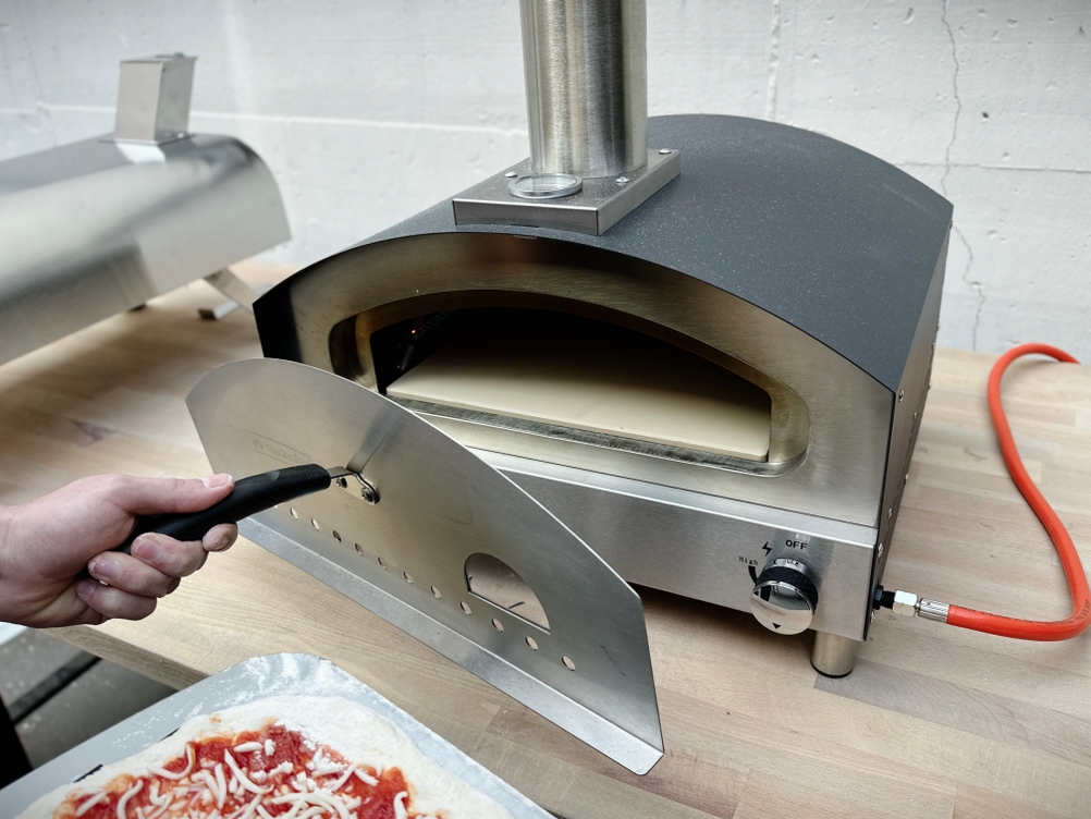 Ein Nutzer öffnet die Metalltür des Heidenfeld KY019G mit einem hitzebeständigen Griff. Die Pizza liegt schon bereit auf der Schaufel.