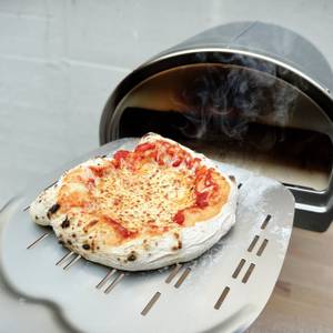 Eine fertig gebackene Pizza wird auf einer gelochten Pizzaschaufel aus dem Gozney Pizzaofen entnommen. Dampf steigt sichtbar auf.