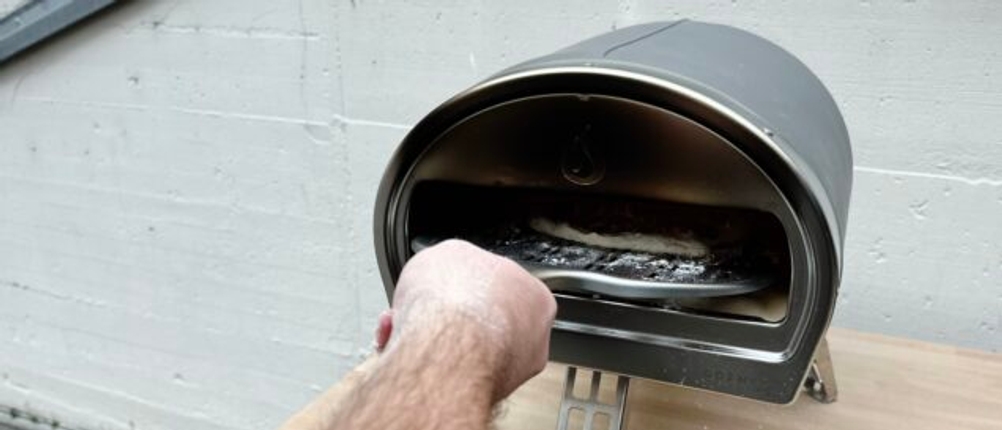 Eine belegte Pizza wird mit einer Schaufel in den Gozney Pizzaofen geschoben. Der Teig ist roh und mit Käse bestreut.