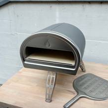 Der Gozney Roccbox Pizzaofen steht aufgebaut auf einem Holztisch. Daneben liegt eine gelochte Edelstahlschaufel.