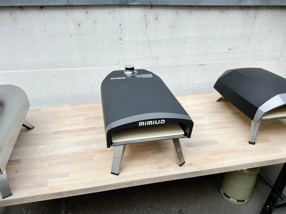 Der Mimiuo-Pizzaofen steht auf einem Holztisch zwischen zwei weiteren Pizzaöfen. Die Beine sind eingeklappt.