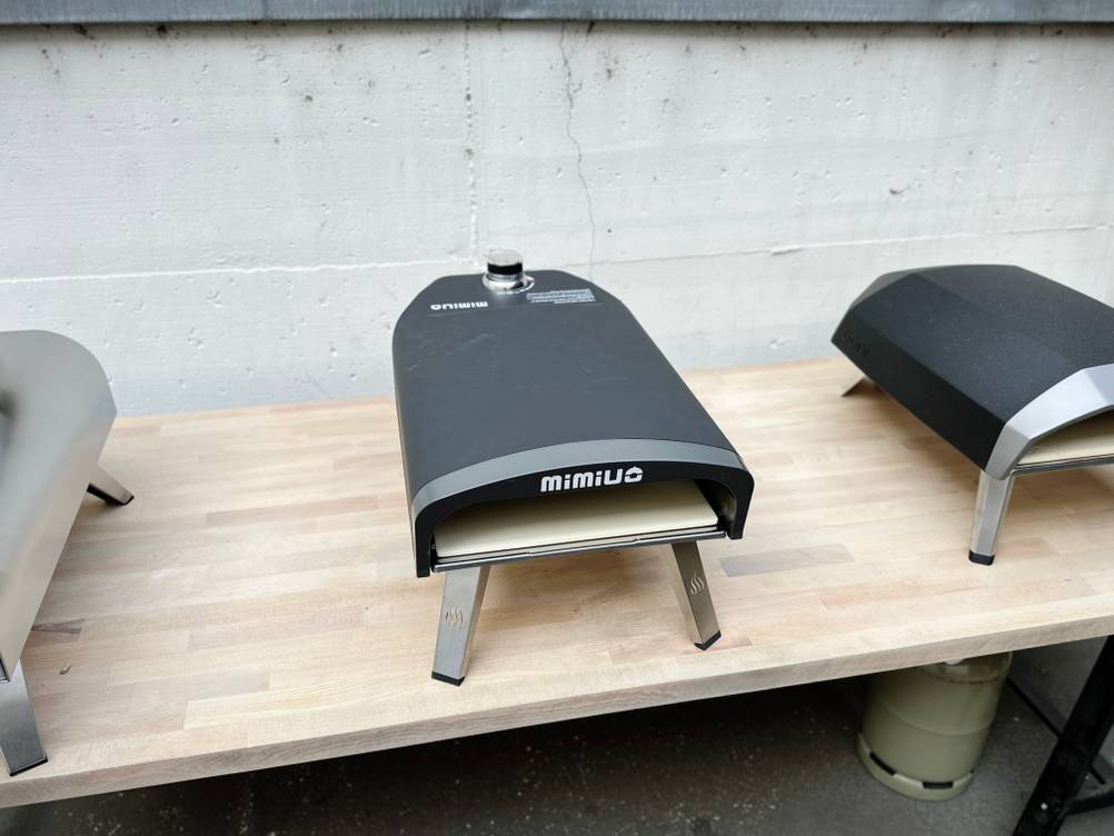 Der Mimiuo-Pizzaofen steht auf einem Holztisch zwischen zwei weiteren Pizzaöfen. Die Beine sind eingeklappt.
