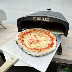 Eine frisch gebackene Pizza liegt auf einer Pizzaschaufel direkt vor dem geöffneten Mimiuo-Pizzaofen.