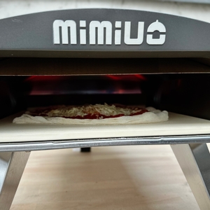 Eine belegte Pizza liegt auf dem Pizzastein im heißen Mimiuo-Pizzaofen. Der Teig beginnt zu backen.