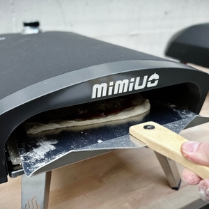 Ein Pizzateig mit Belag wird mit einer Holzgriff-Schaufel in den Mimiuo-Pizzaofen geschoben.