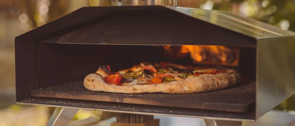 Pizzaofen im Test: Mobiler Pizzaofen steht auf einer Unterlage, im Ofen ist eine Pizza und unter dem Pizzaofen liegt eine Bürste.