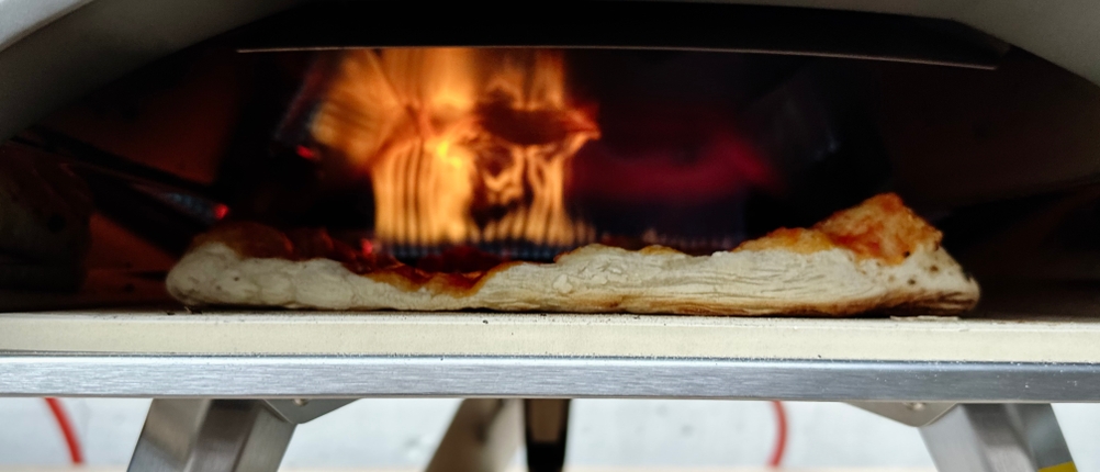 Die Flamme im Ooni Koda 12 ist im Hintergrund sichtbar, während eine Pizza auf dem Stein backt. Der Boden hebt sich durch die Hitze leicht an.