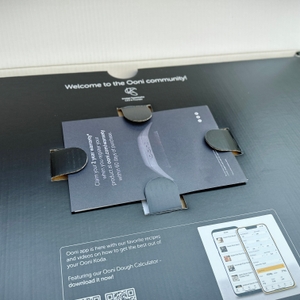 Die Verpackung des Ooni Koda 12 ist geöffnet. Im Inneren liegt eine Broschüre mit QR-Code zur App und Rezeptanleitung.