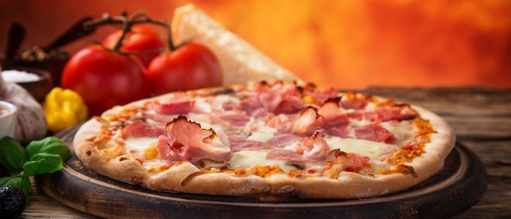 Eine Pizza mit Schinken liegt auf einem Teller. Im Hintergrund sind Tomaten, italienischer Hartkäse und weitere Zutaten zu sehen.