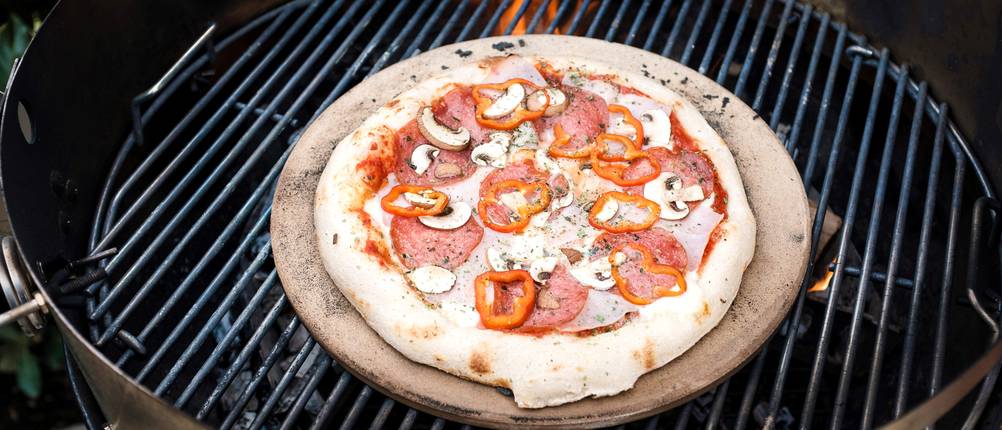 Eine Pizza liegt auf einem Pizzastein im Kugelgrill.