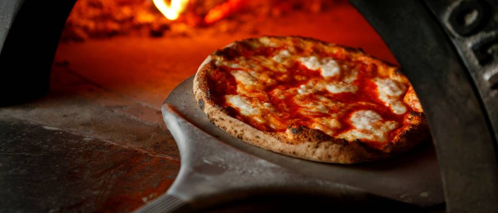 Eine Pizza wird mit einer Pizzaschaufel aus Metall in einen Pizzaofen geschoben.