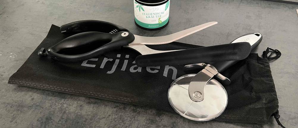Auf einem Säckchen liegt eine Pizzaschere im Test und ein Pizzaschneider vor einer Dose mit Kräutern.
