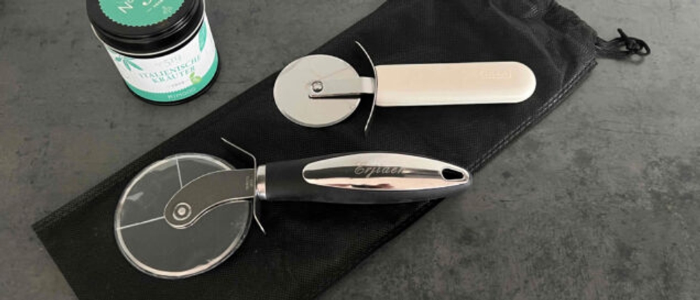 Auf einer dunklen Fläche liegen zwei Pizzaschneider im Test dahinter eine Dose mit Kräutern.