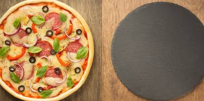 Pizza neben Pizzastein und Zutaten auf Holzfläche.