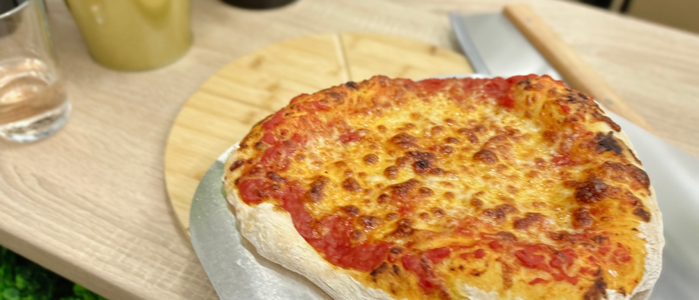 Eine fertig gebackene Pizza liegt auf einer Metallschaufel vor einem Holzbrett. Im Hintergrund stehen Pflanzen und ein Wasserglas.