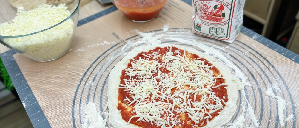 Ein belegter Pizzateig mit Tomatensoße und Käse liegt auf einer Backmatte. Neben dem Teig stehen Mehl, Käse und Sauce bereit.
