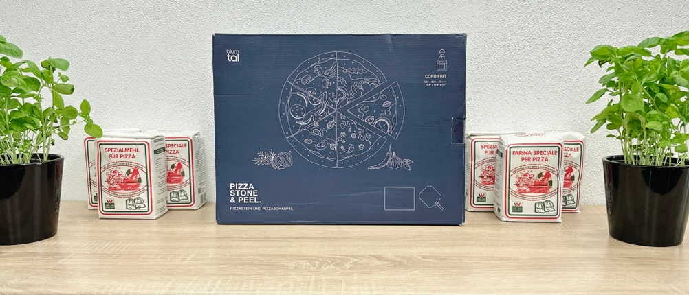 Die Verpackung des Blumtal Pizzaofen-Kits steht mittig auf einem Tisch. Links und rechts stehen Mehlpackungen und Kräutertöpfe.