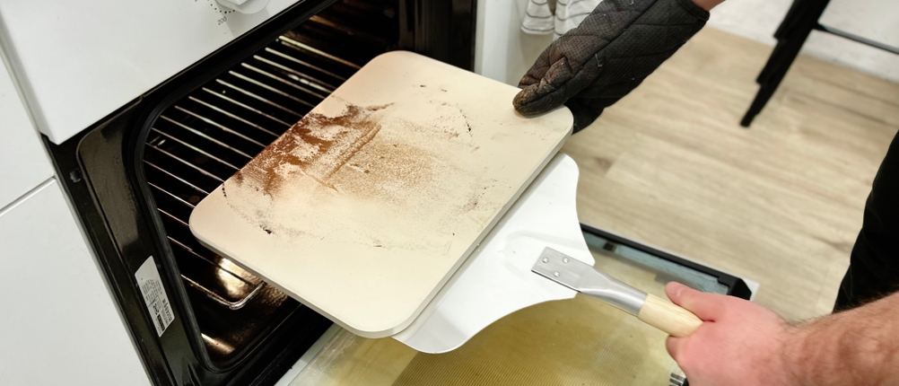 Ein benutzter Pizzastein von Blumtal wird mit einer Metallschaufel aus dem Ofen geholt. Der Stein zeigt Backrückstände und ist leicht gebräunt.