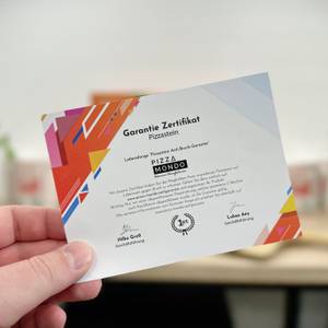Eine Hand hält das Garantie-Zertifikat des Pizza Mondo Pizzasteins in die Kamera. Im Hintergrund sind Pflanzen unscharf zu sehen.