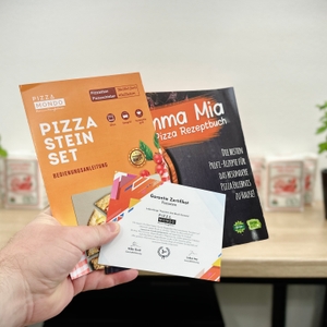 Eine Hand hält Anleitung, Rezeptheft und Garantie-Zertifikat des Pizza Mondo Pizzastein Sets in die Kamera. Im Hintergrund stehen Basilikumpflanzen.