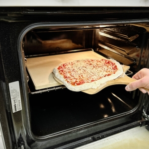 Eine Pizza wird mit einem Holzschieber in den Backofen auf den Pizzastein geschoben. Eine Hand führt den Schieber vorsichtig.