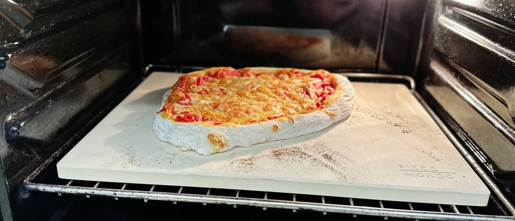 Im Backofen liegt eine fast fertig gebackene Pizza auf einem weißen Pizzastein mit der Aufschrift Pizza Mondo.