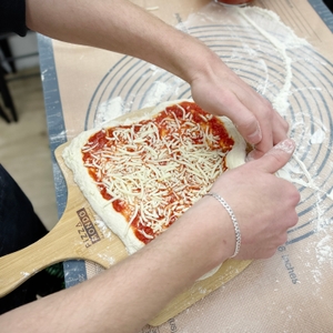 Ein belegter Pizzateig mit Tomatensoße und Käse liegt auf einer Backmatte. Neben dem Teig stehen Mehl, Käse und Sauce bereit.