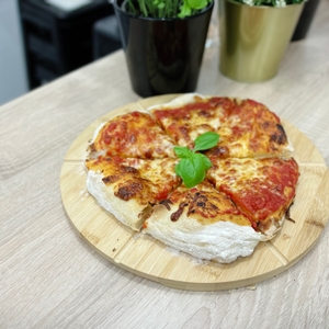 Auf einem Holzbrett liegt eine aufgeschnittene Pizza mit Basilikum in der Mitte. Im Hintergrund stehen zwei Blumentöpfe auf der Küchenarbeitsplatte.