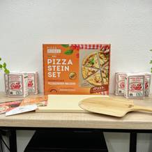 Das Pizza Mondo Pizzastein-Set liegt mit Zubehör auf einem Tisch. Verpackung, Schieber, Zertifikate und Pizzamehl sind sichtbar.
