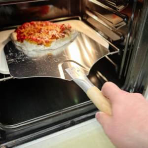 Eine belegte Pizza wird mit einer Metallschaufel in den Backofen auf einen Pizzastein geschoben.
