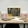 Das komplette Set von Locbird mit Pizzastein, Schieber, Rezeptbuch und Mehl steht auf einem Tisch. Kräutertöpfe rahmen das Bild ein.