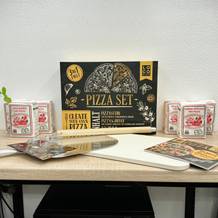 Das komplette Set von Locbird mit Pizzastein, Schieber, Rezeptbuch und Mehl steht auf einem Tisch. Kräutertöpfe rahmen das Bild ein.