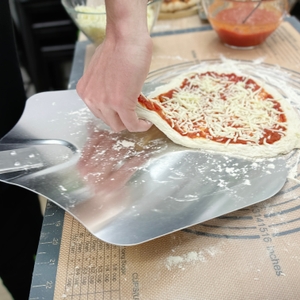 Ein belegter, roher Pizzateig wird auf einer bemehlten Metallschaufel vorbereitet. Eine Hand hebt den Rand leicht an.