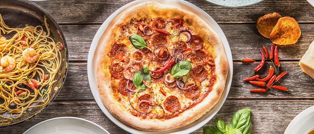 Pizzateller-Test: Blick auf eine Salamipizza von oben, umrahmt von Basilikum, Chili und weiteren Tellern mit Pastagerichten.