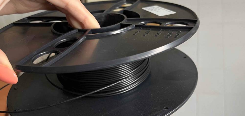 Hand hält eine getestete 3D-Drucker-Filament-Rolle mit schwarzem Drucker-Faden vor einen hellen Hintergrund.