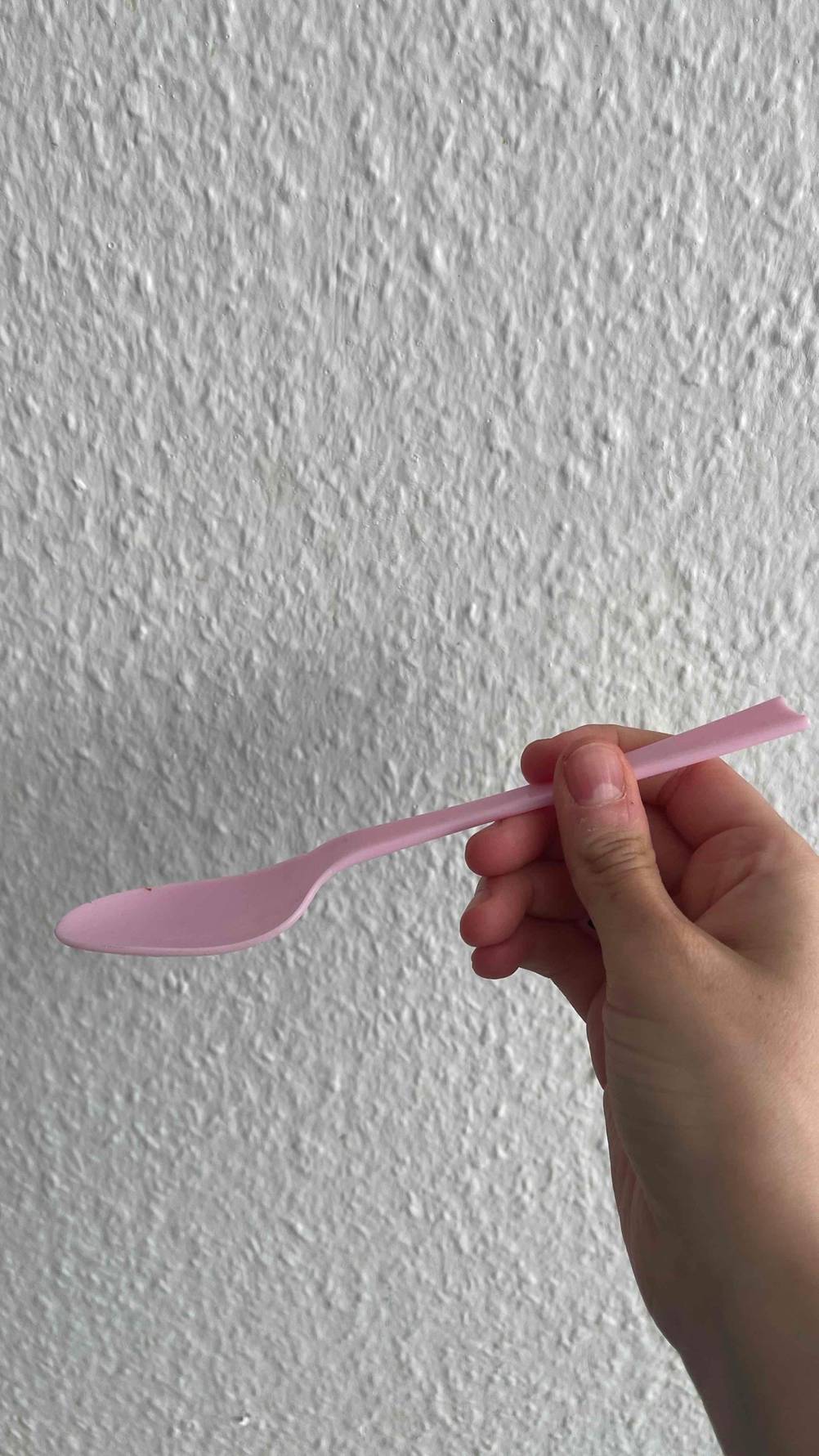 Ein getesteter Plastiklöffel von der Seite von einer Hand vor einer helle Wand gehalten.