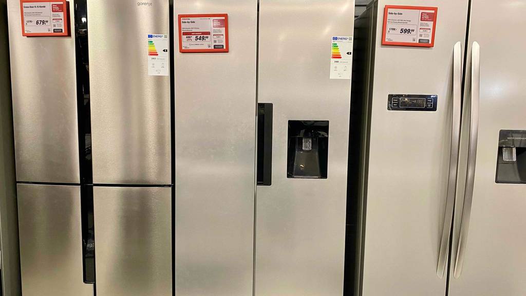 Side-by-Side-Kühlschrank-Test: Ansicht von vorn auf ein silberfarbenes Modell inklusive schwarzem Wasserspender, Handgriff und Preisschild zwischen weiteren Modellen in einem Verkaufsbereich.