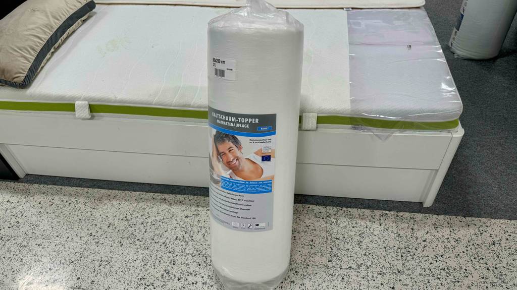 Ein Kaltschaum Matratzen-Topper im Test steht in Kunststoff eingerollt vor einer auf einem Bettgestell ausgestellten Matratze.