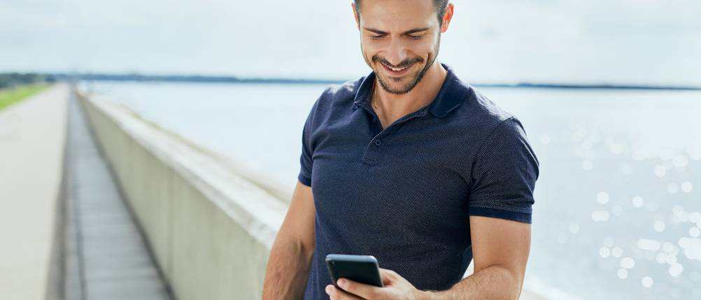 Poloshirt für Herren im Test: Mann trägt ein Poloshirt, steht am Meer und schaut auf das Handy.