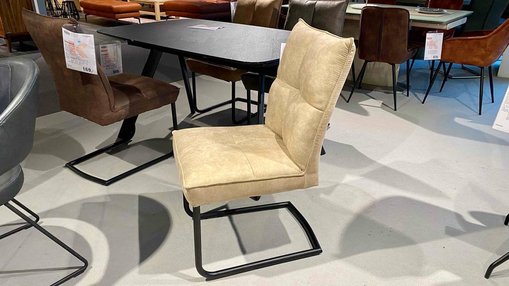 Im Test: Schwingstuhl in Braun und in Beige in einem Einkaufsbereich zwischen anderen Stühlen und Tischen.