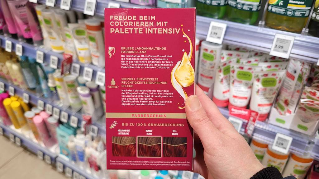 Eine Hand hält eine Packung, mit Fokus auf die Produktbeschreibung, einer getesteten roten Haarfarbe vor einem Drogeriemarktregal.